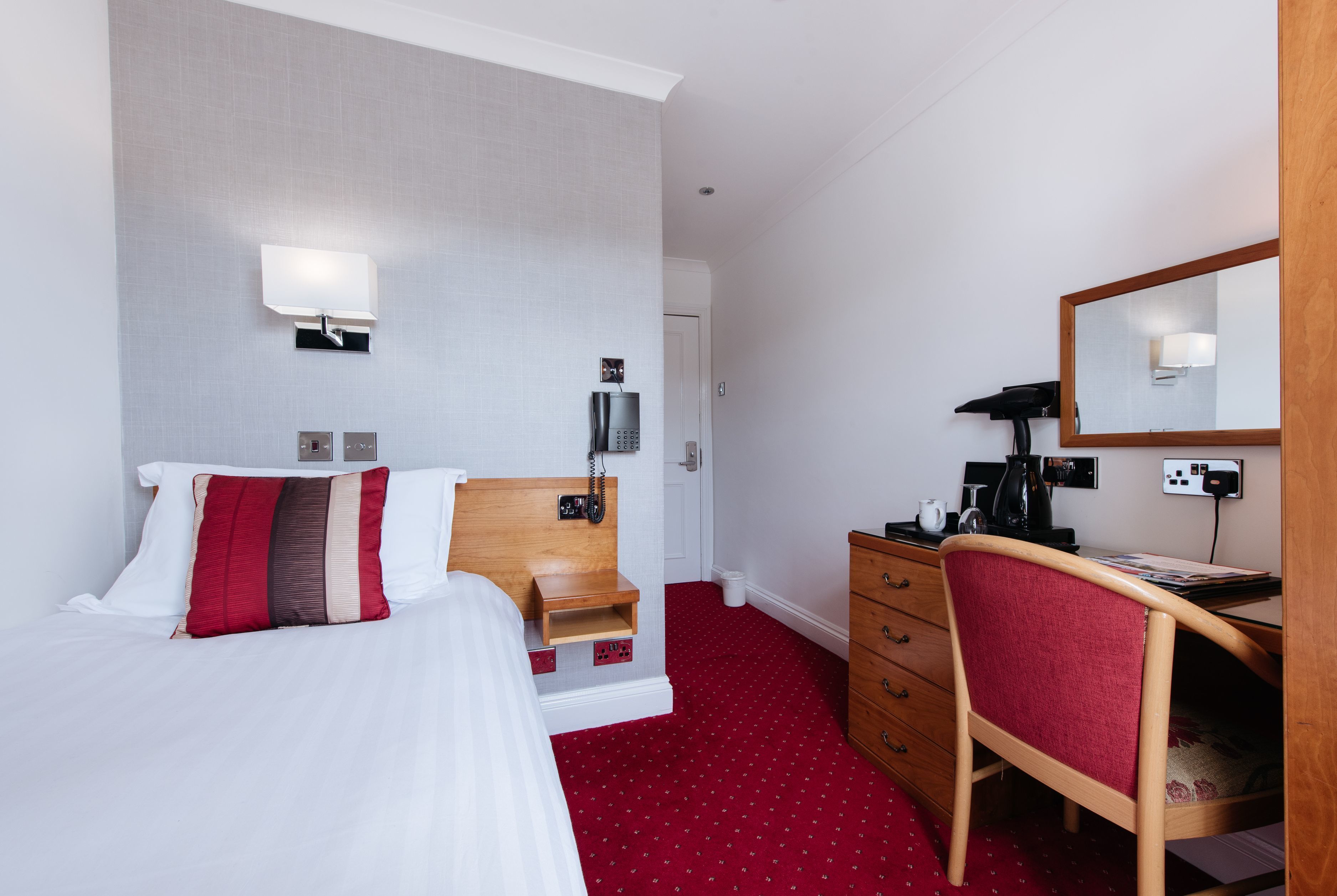 Una camera d'albergo con un letto, una scrivania con una sedia, uno specchio e un tappeto rosso.
