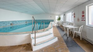 Una piscina con escalones de acceso, rodeada de sillas y mesas blancas, y carteles que indican que no se permite bucear ni nadar sin supervisión.