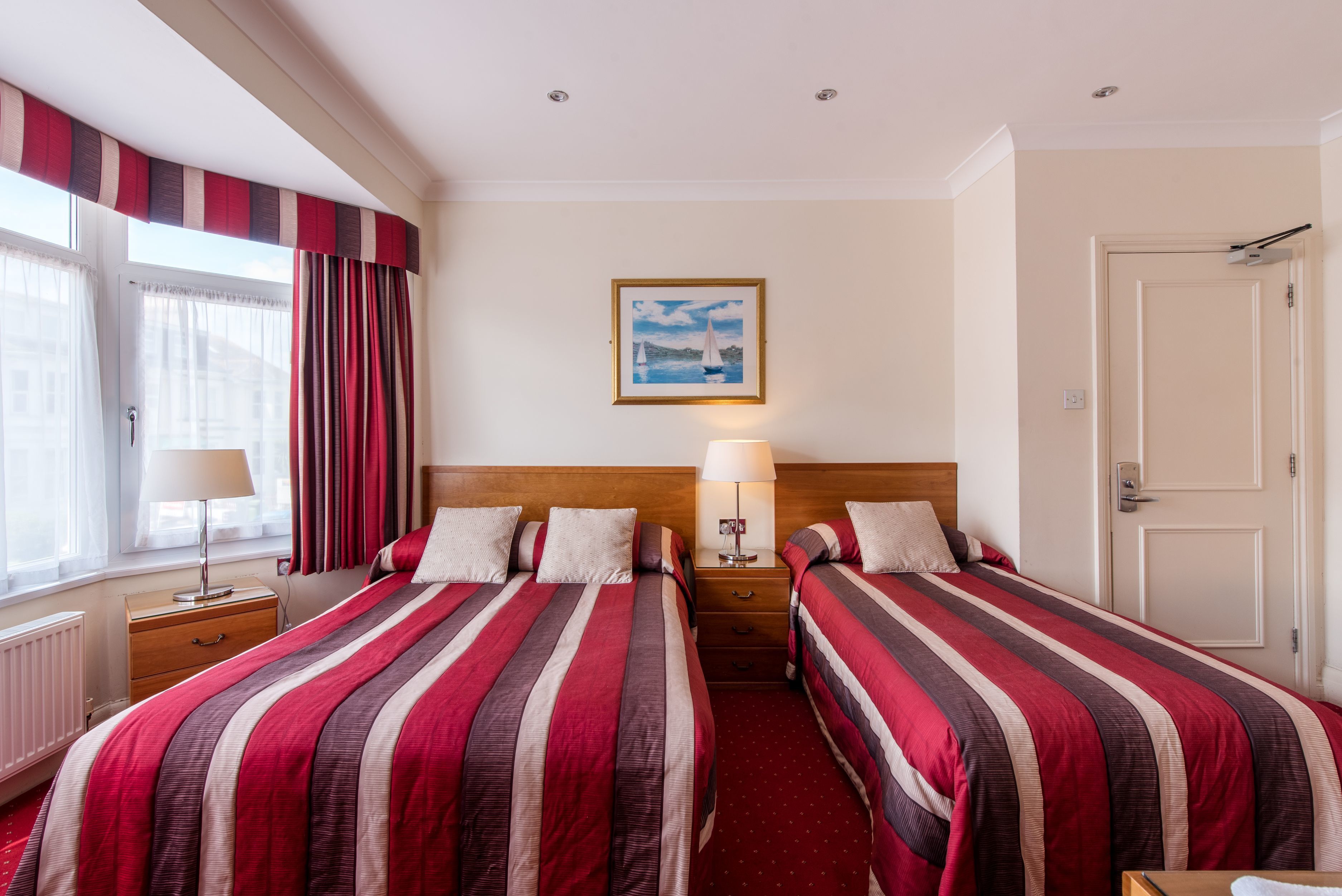A hotel room with two beds, a wooden headboard, a nightstand with a lamp, a window with curtains, and a painting on the wall.