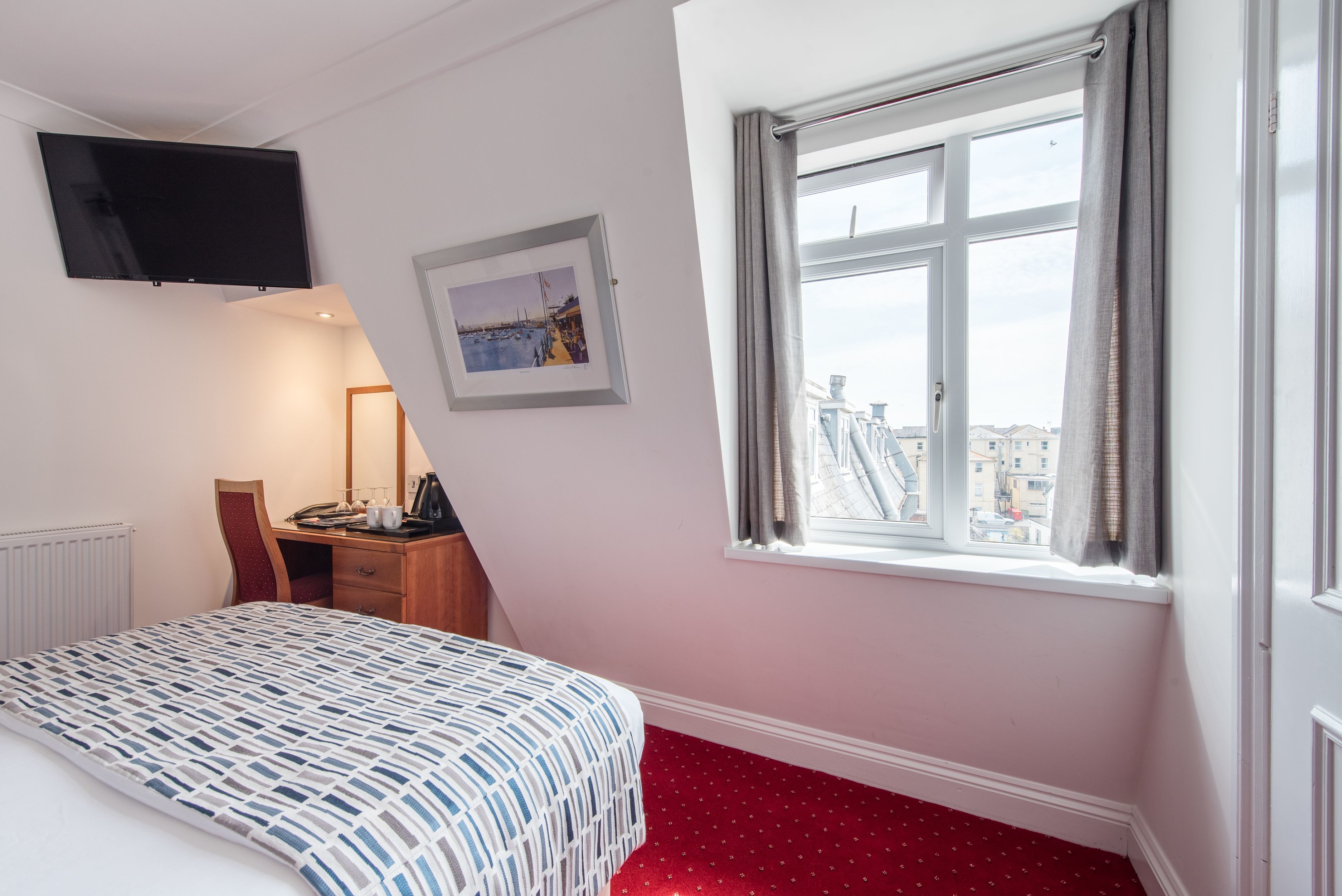 A hotel room with a bed, a desk, a flat-screen TV mounted on the wall, a framed picture, and a window with a view of buildings.