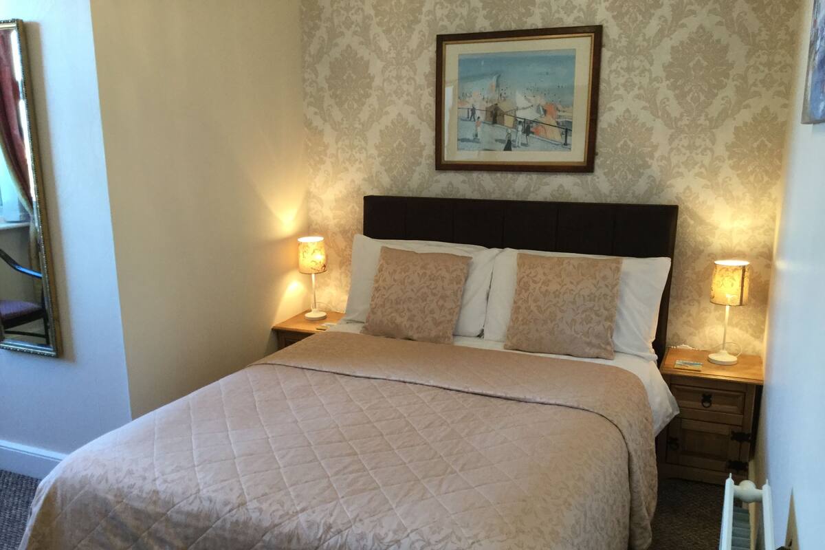 A neatly made bed with a quilted cover, two bedside lamps, a framed picture on the wall, and a patterned wallpaper.