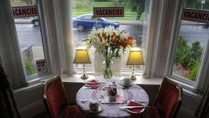 A dining area with a table set for two, two lamps, a vase with flowers, and a view of the street with 'VACANCIES' signs. - Balmoral (Skegness)