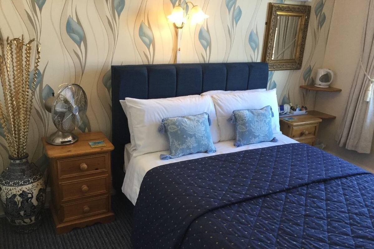 A neatly made bed with a dark blue quilt, a wooden nightstand with a fan, and a decorative vase.