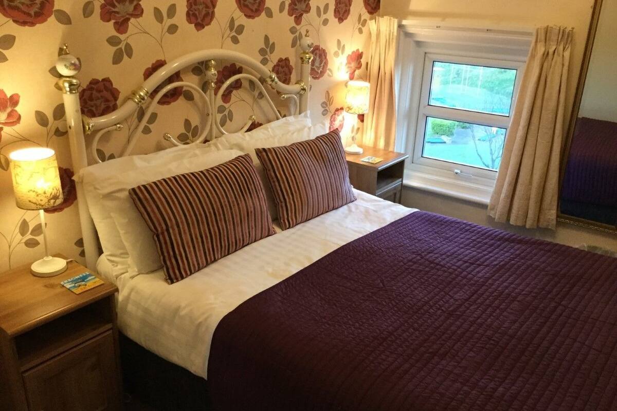 A neatly made bed with a striped pillow and a dark quilt, a wooden nightstand with a lamp, and a window with curtains.