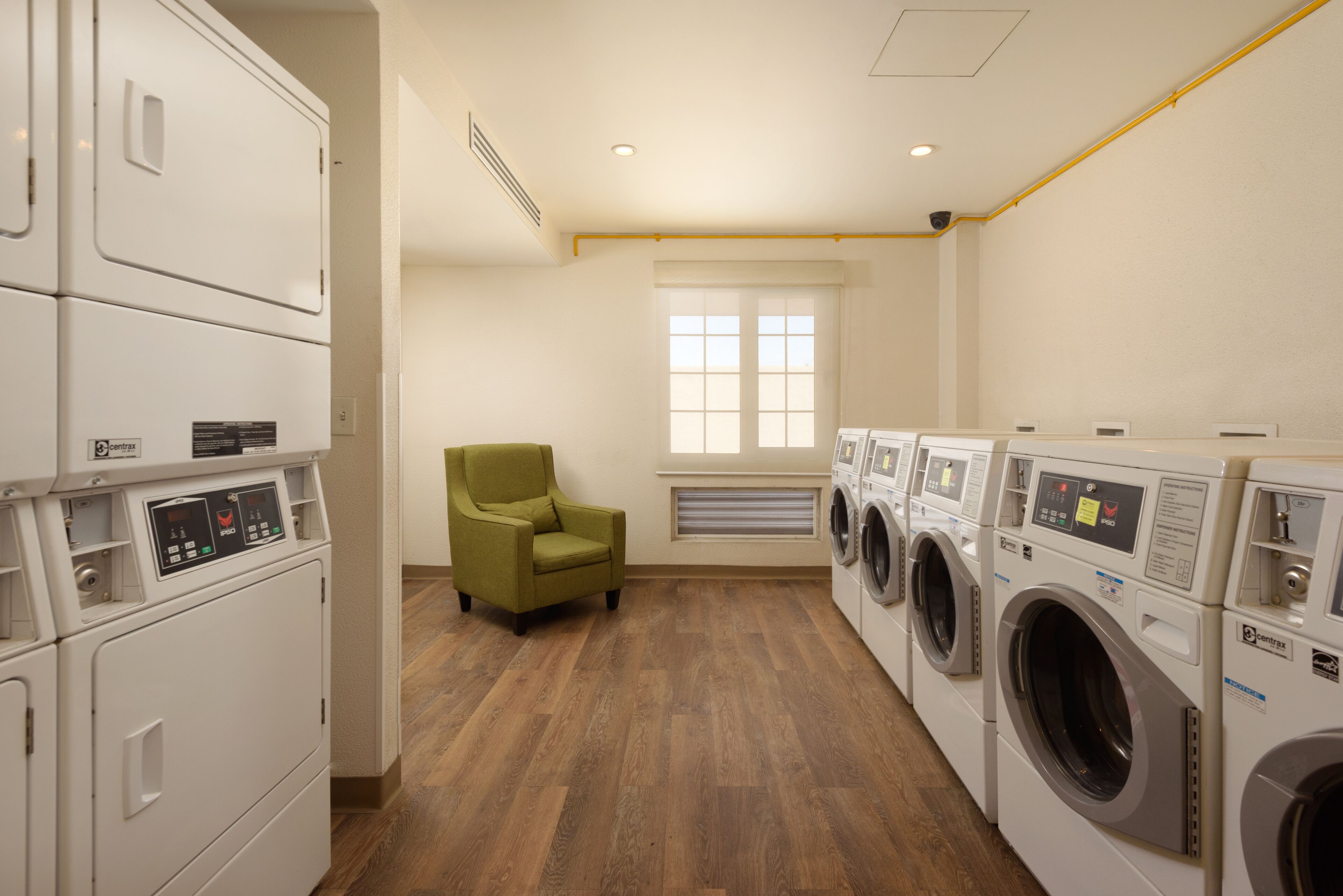 A laundry room with stacked washers and dryers, a green chair, and a window with blinds.