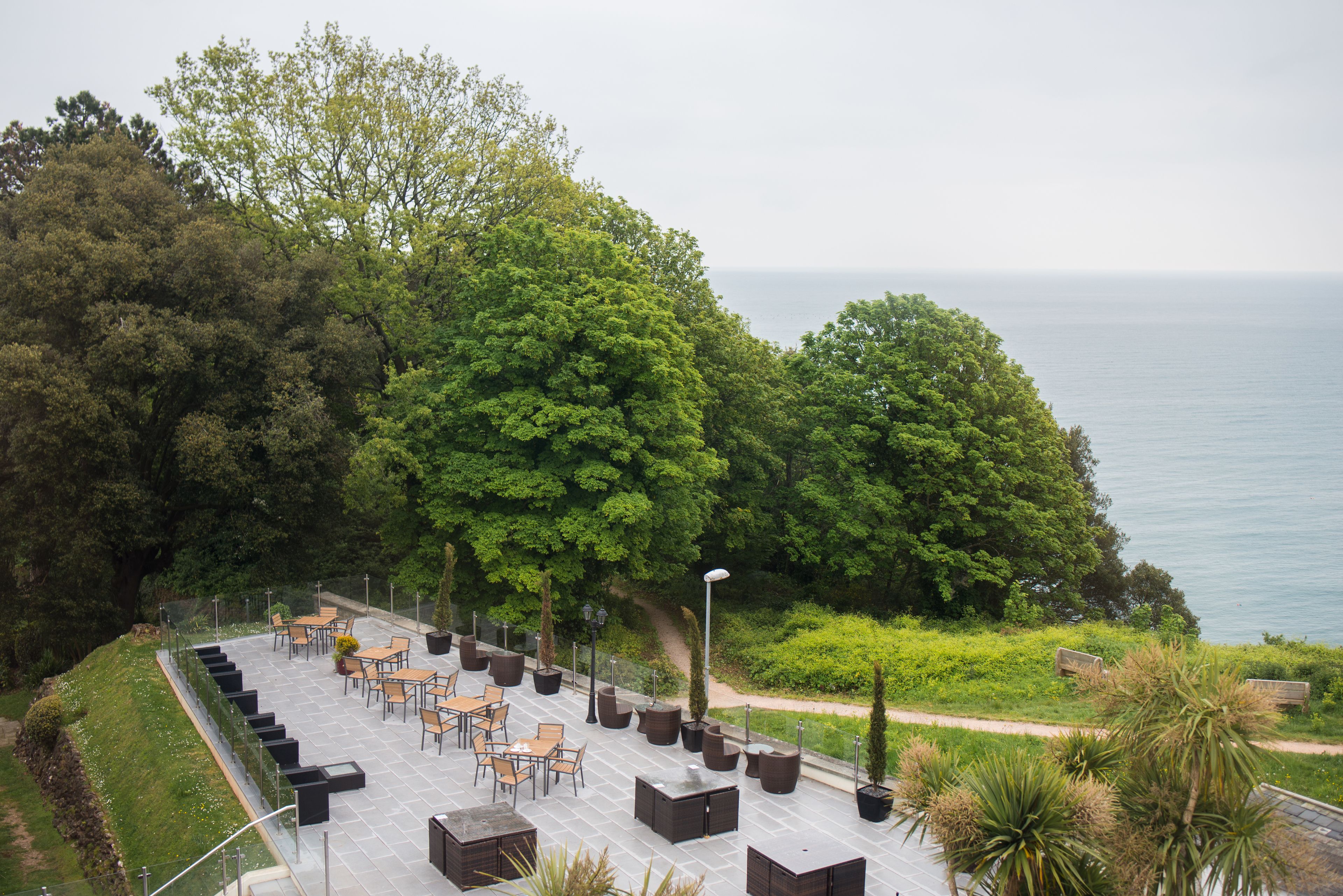 An outdoor seating area with tables and chairs, surrounded by greenery and overlooking a body of water.