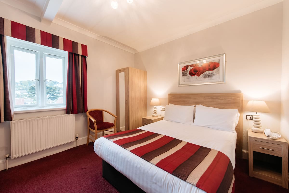 A hotel room with a large bed, two bedside tables with lamps, a chair, a radiator, a window with curtains, and a framed picture on the wall.