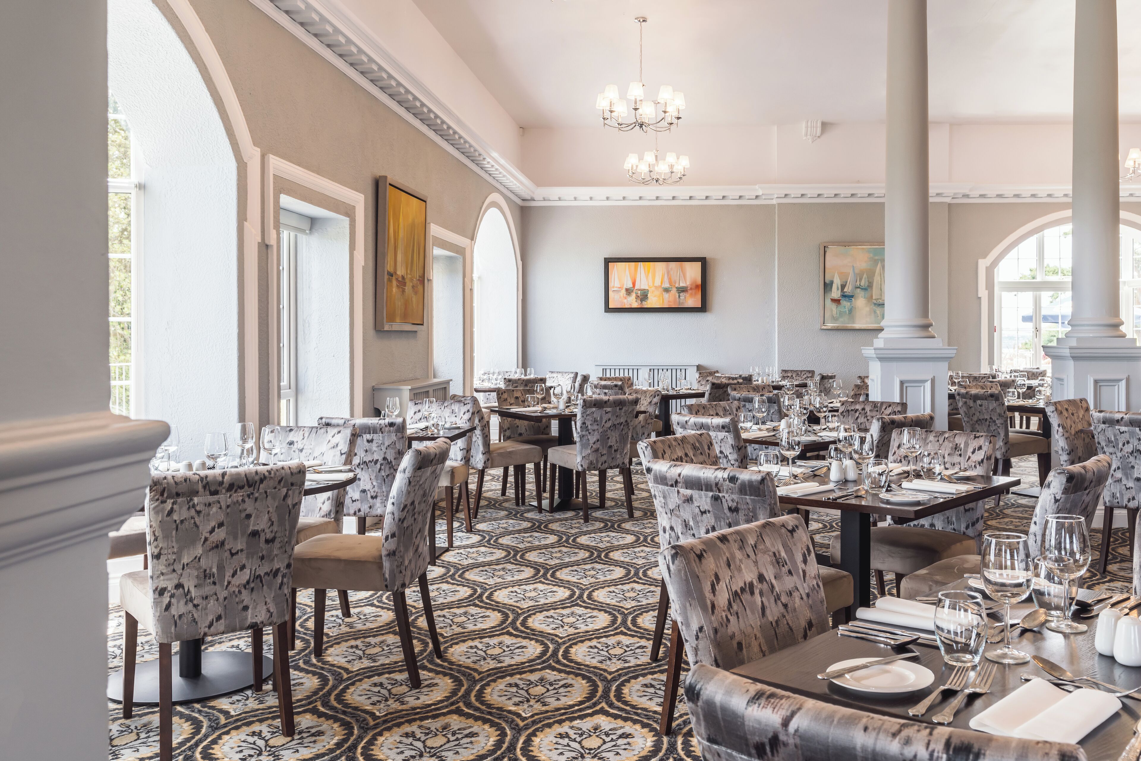 A spacious dining area with multiple tables and chairs, large windows, and a patterned carpet.