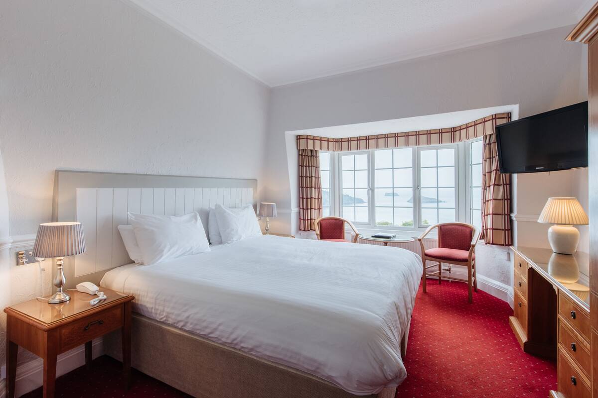 A hotel room with a large bed, a desk with a lamp, a chair, a TV, and a window with curtains.
