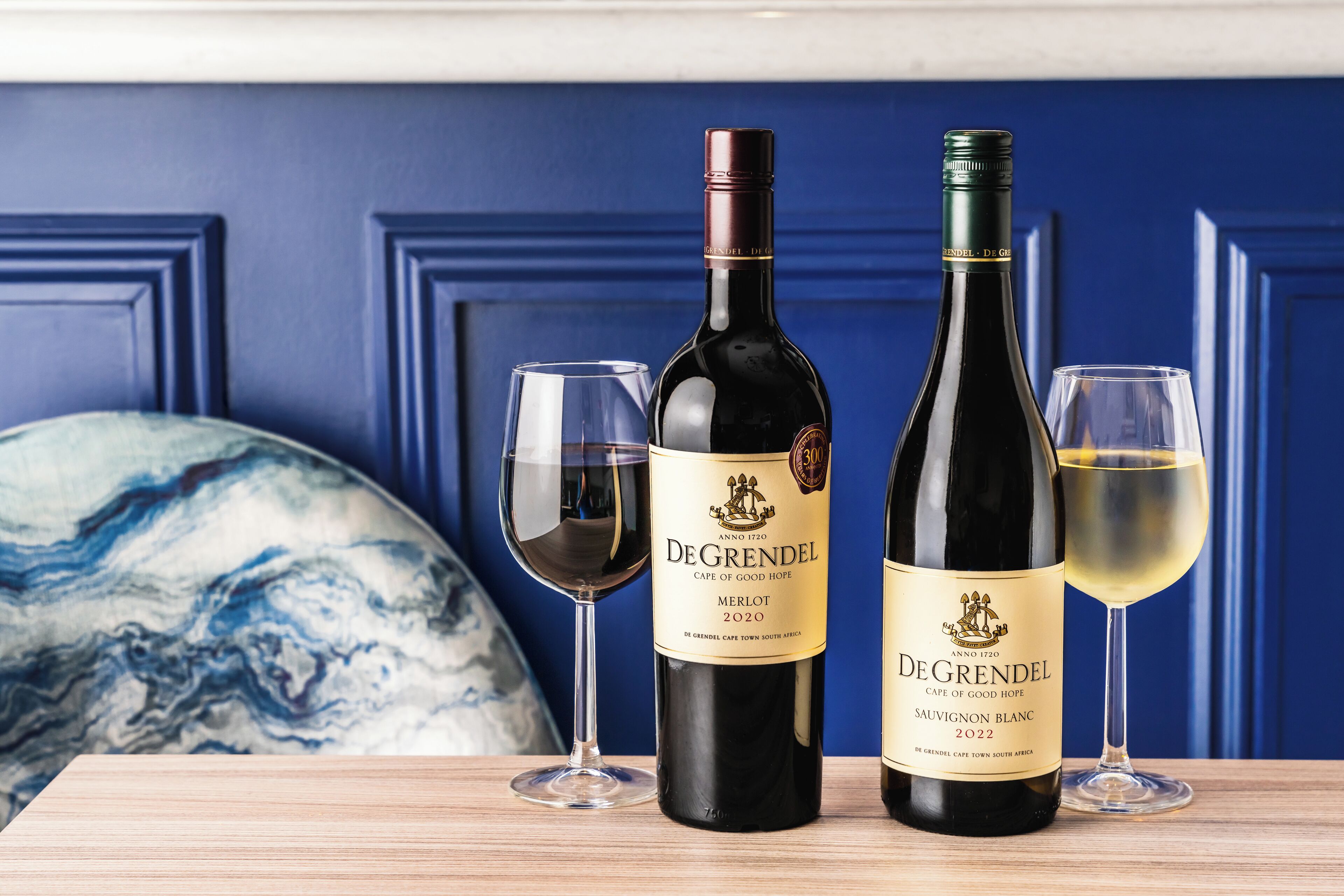 Two bottles of DeGrendels wine, one Merlot and one Sauvignon Blanc, with two glasses of wine on a wooden table against a blue wall.