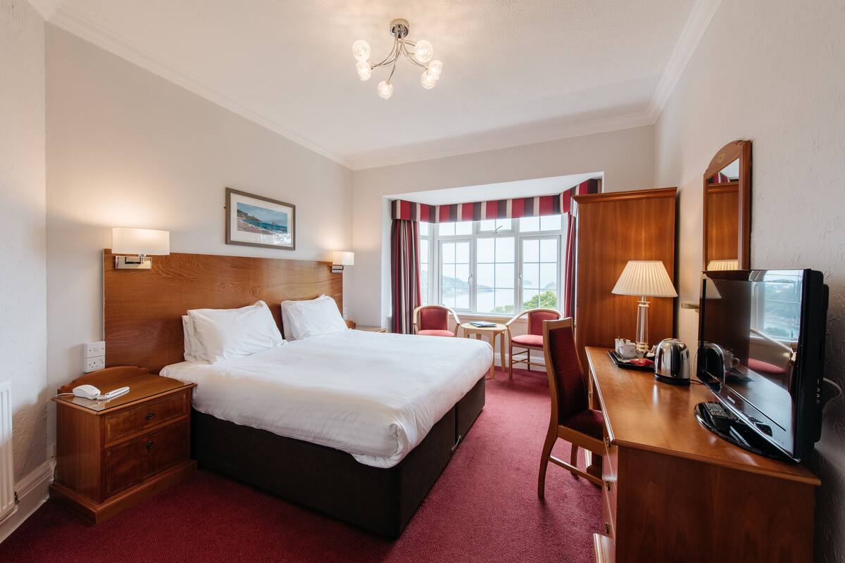 A hotel room with a large bed, a desk with a television, a chair, and a window with curtains.