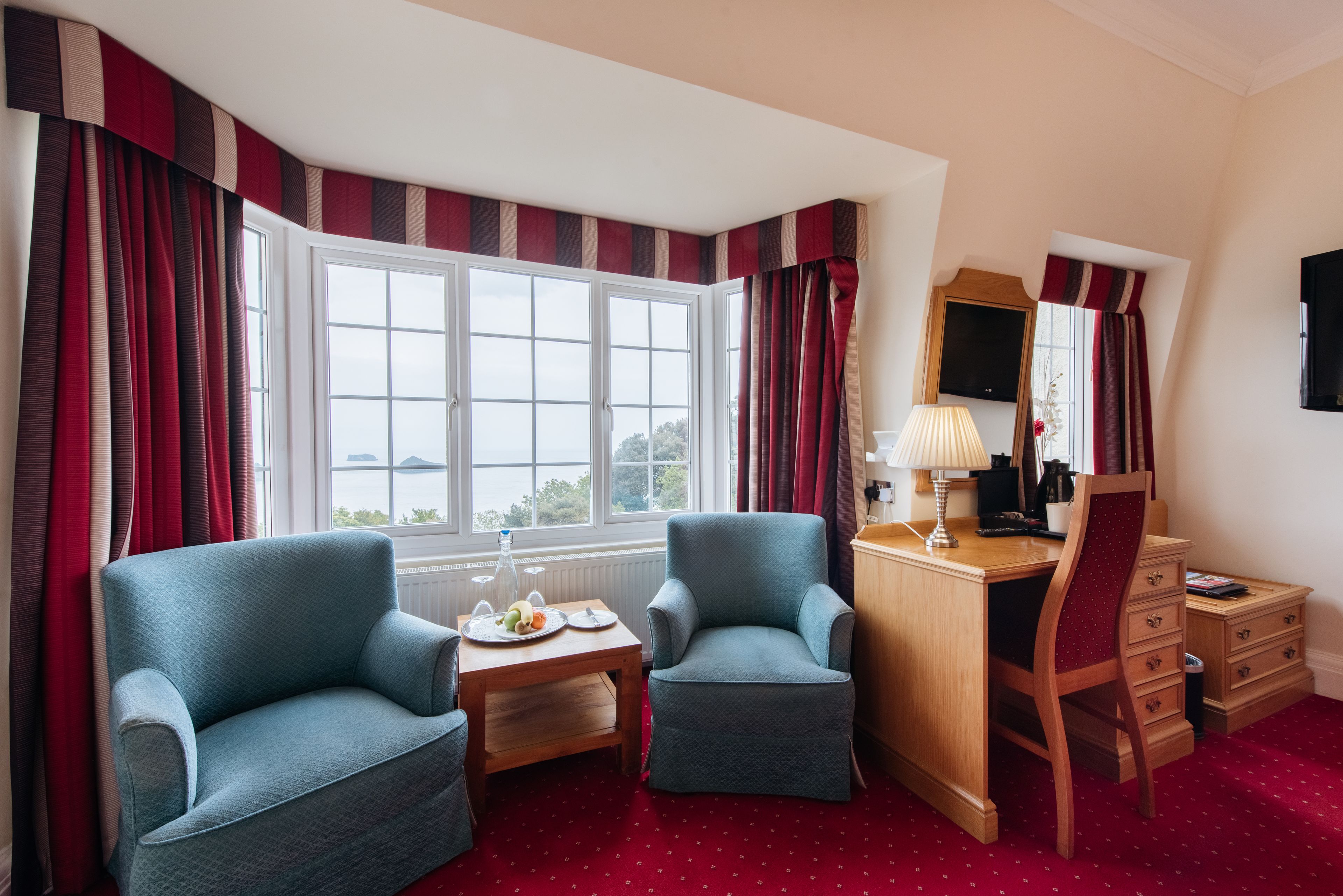Quarto de hotel com duas poltronas, uma secretária de madeira, televisão e janela com cortinas.