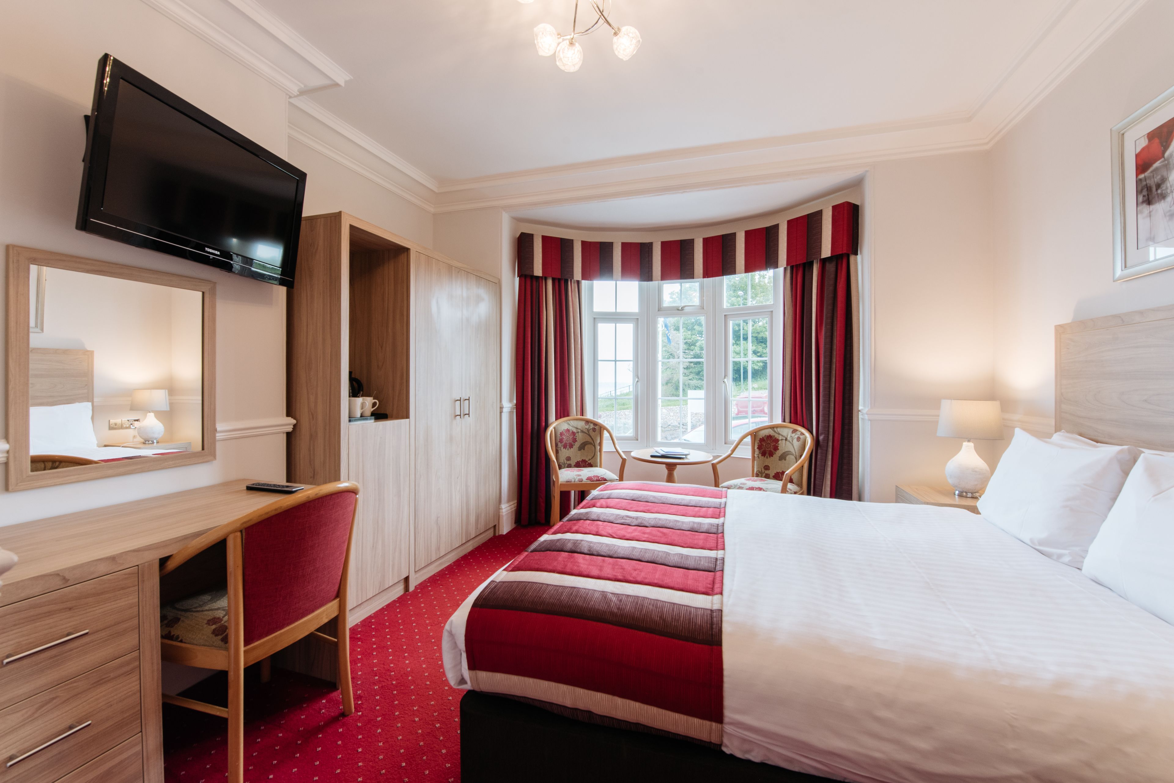 A hotel room with a bed, a desk, a TV, a window with curtains, and a view of greenery outside.