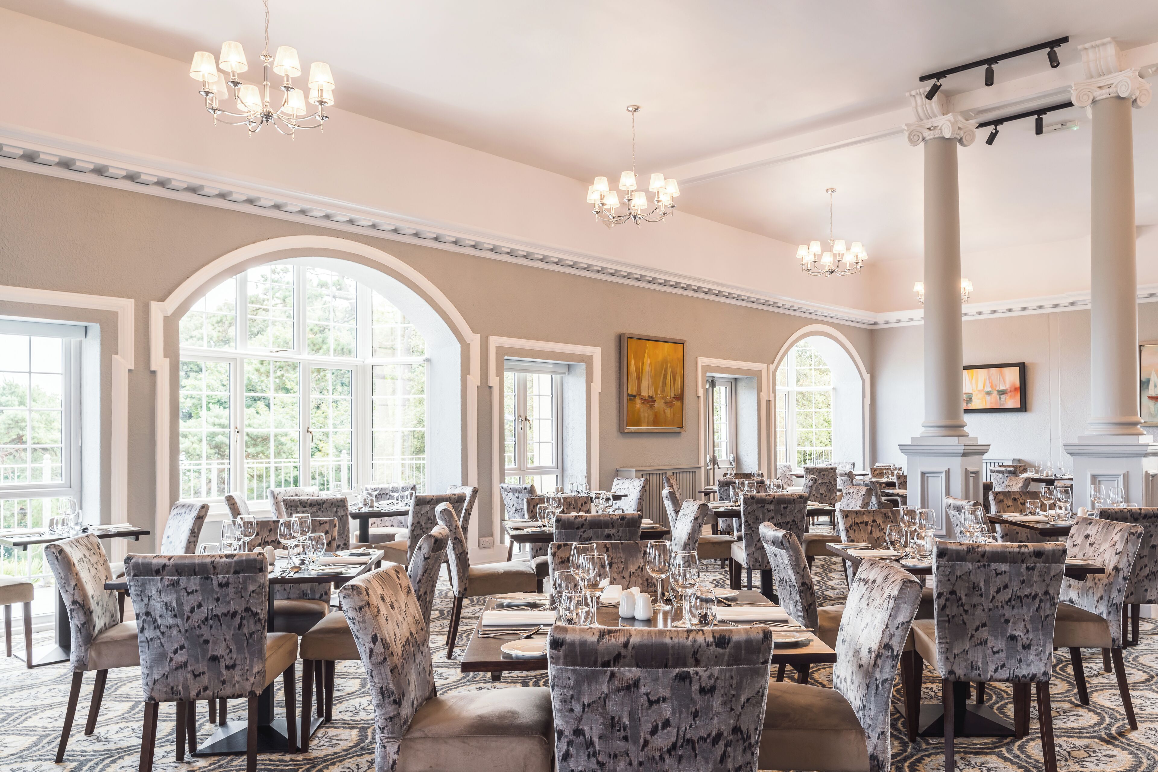 A spacious dining area with high ceilings, chandeliers, and large windows allowing natural light. Tables are set with chairs, and the room features classic architectural details.