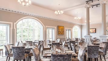 A spacious dining area with high ceilings, chandeliers, and large windows allowing natural light. Tables are set with chairs, and the room features classic architectural details.