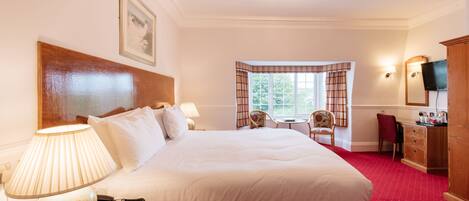 A hotel room with a large bed, a desk with a lamp, a chair, a television, and a window with curtains.