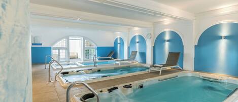 En stor indendørs swimmingpool med flere jacuzzier, liggestole og uhindret udsigt til det fri gennem store vinduer.