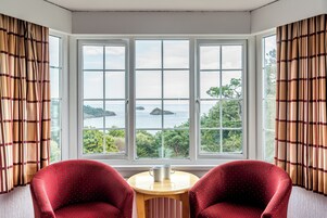 Quarto com uma grande janela em arco, duas cadeiras vermelhas e uma pequena mesa com duas canecas brancas.