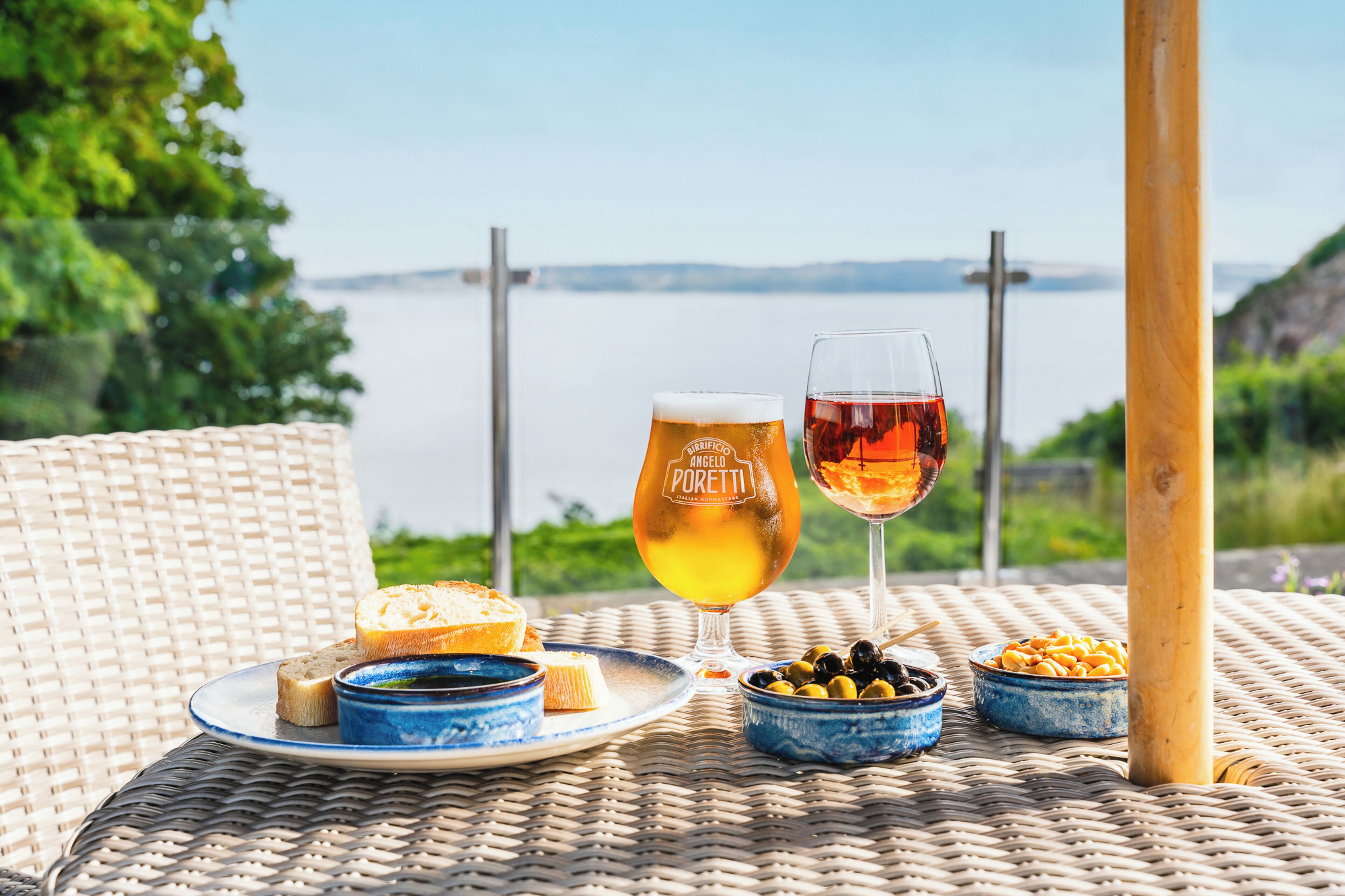 Une table en osier, avec un verre de bière, un verre de vin rouge et une assiette de pain et d’olives.