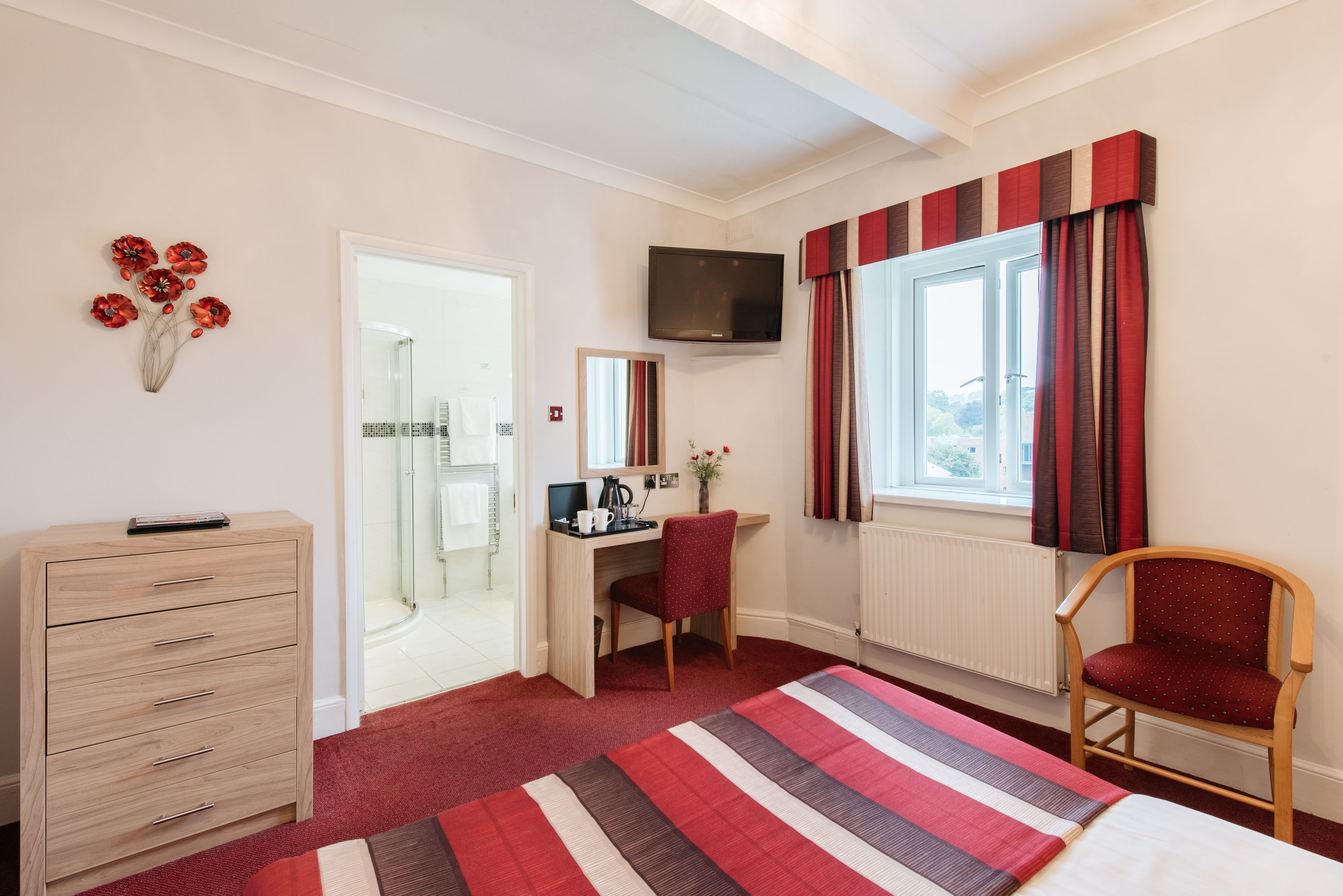 Una camera d'albergo con una scrivania in legno, una sedia, un letto con copriletto a righe, un comò, una TV fissata al muro e una finestra con tende.