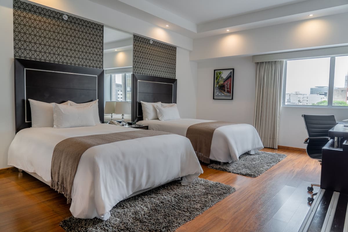 Quarto de hotel com duas camas, uma escrivaninha, uma cadeira e uma janela grande.