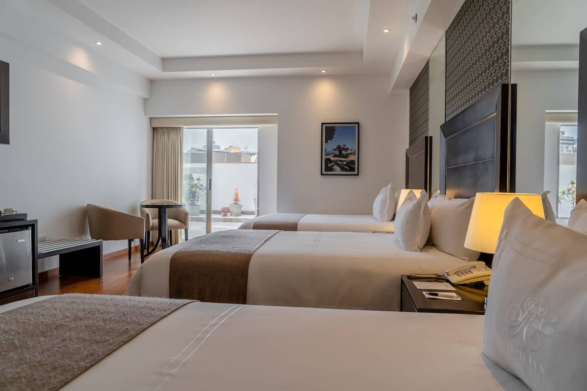 Quarto de hotel com duas camas, uma escrivaninha, uma cadeira, um abajur e uma janela com vista para edifícios.