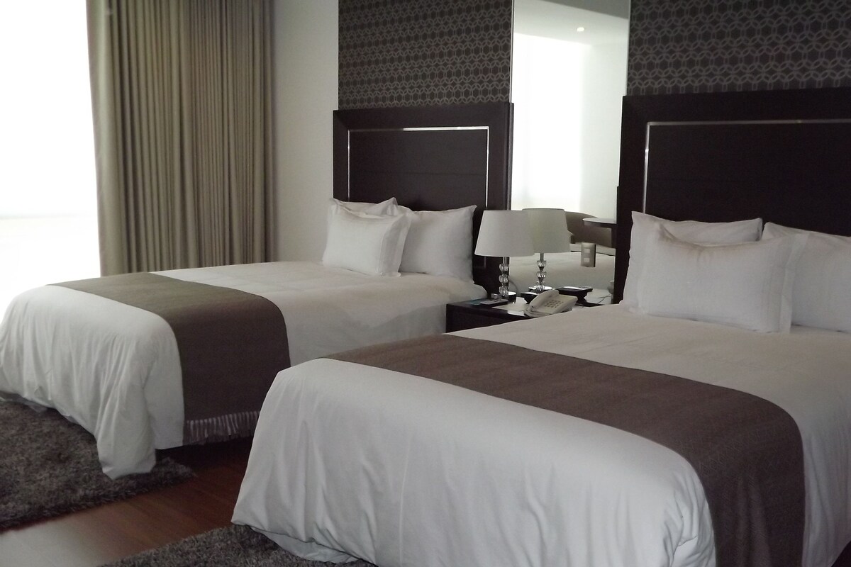 A hotel room with two beds, a nightstand with a lamp, and a mirror.