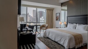 Habitación de hotel con una cama grande, un escritorio con silla, una mesita y vistas a la ciudad a través de amplios ventanales.