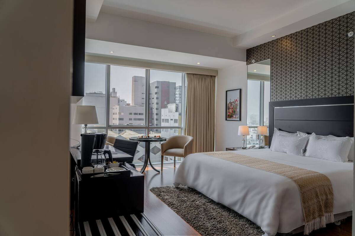 Quarto de hotel com uma cama grande, uma escrivaninha com cadeira, uma mesinha e vista para a cidade por grandes janelas.