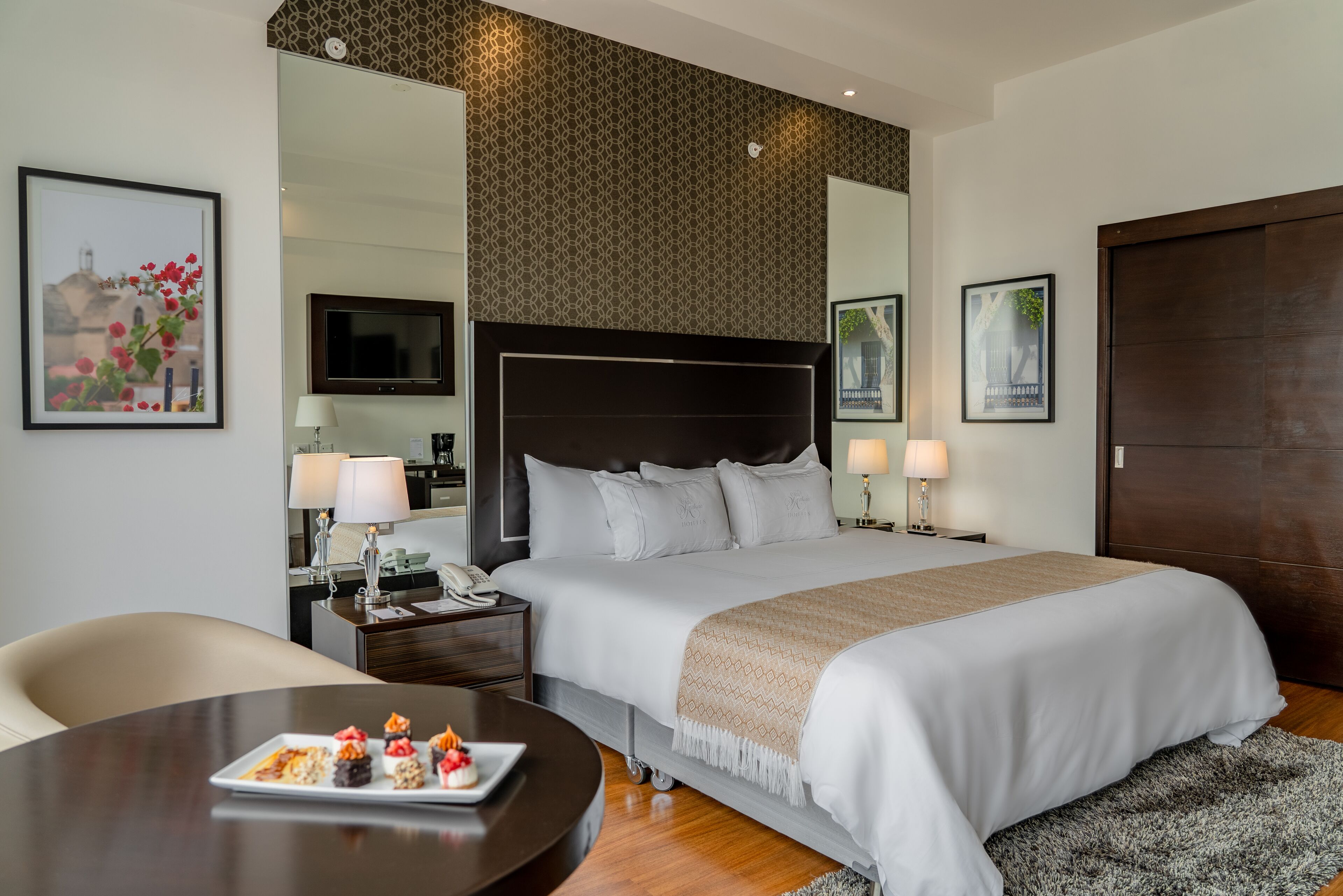 Une chambre d’hôtel avec un grand lit, des tables de chevet avec des lampes, un bureau avec une assiette de nourriture et des œuvres d’art accrochées aux murs.