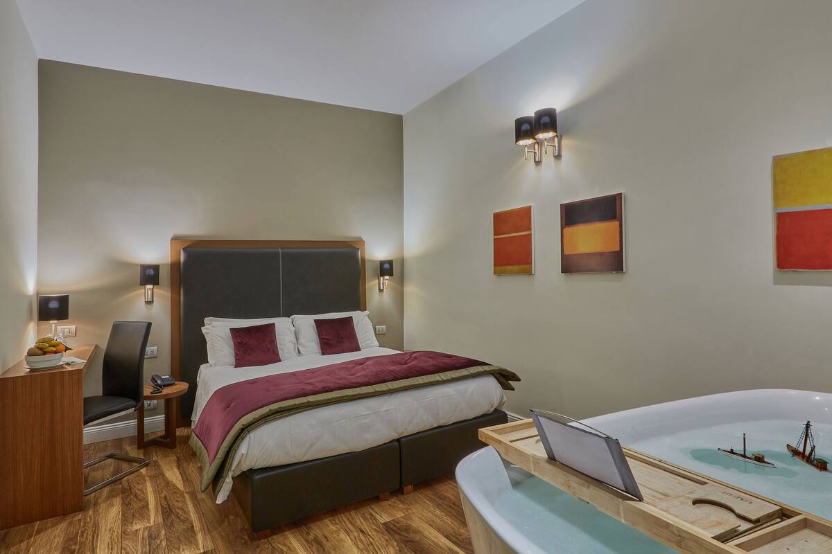 A modern hotel room with a large bed, bedside table, chair, desk, and a bathtub with a ship model.