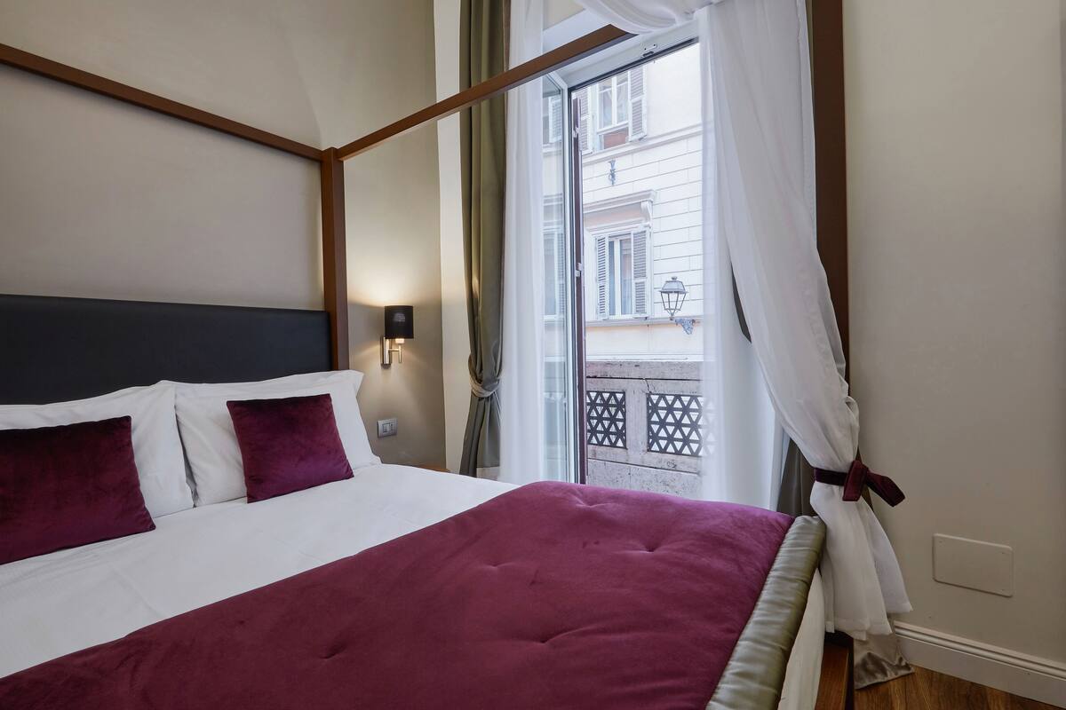 A neatly made bed with a white and maroon bedspread, a dark headboard, and two maroon pillows. A wall-mounted light fixture is visible on the wall.