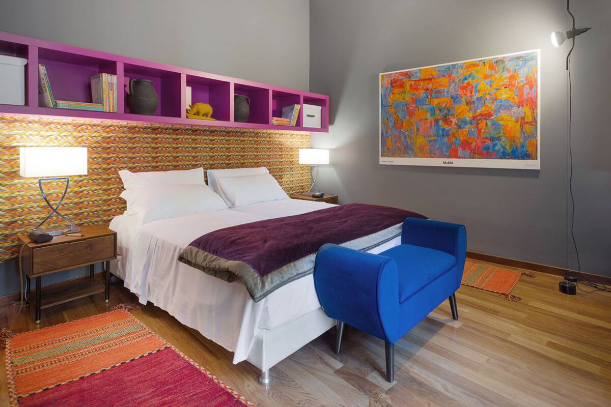 A modern bedroom with a bed, a blue chair, a colorful rug, and a shelf with books and decorative items.