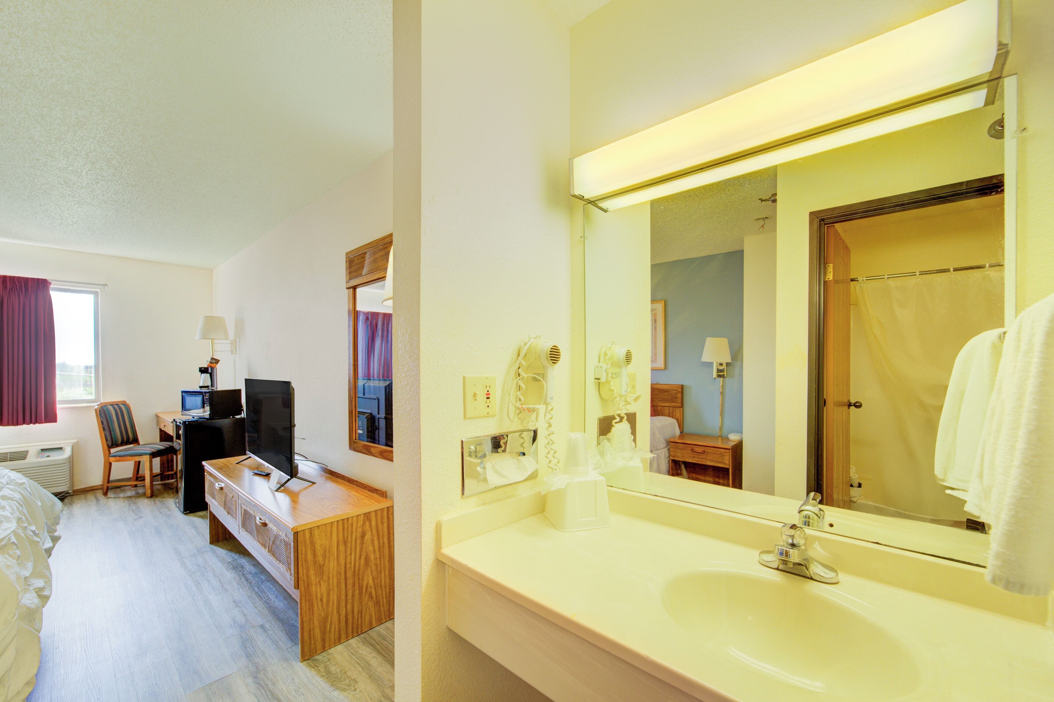 Una habitación de hotel con cama, escritorio, televisor y baño con lavabo y espejo.