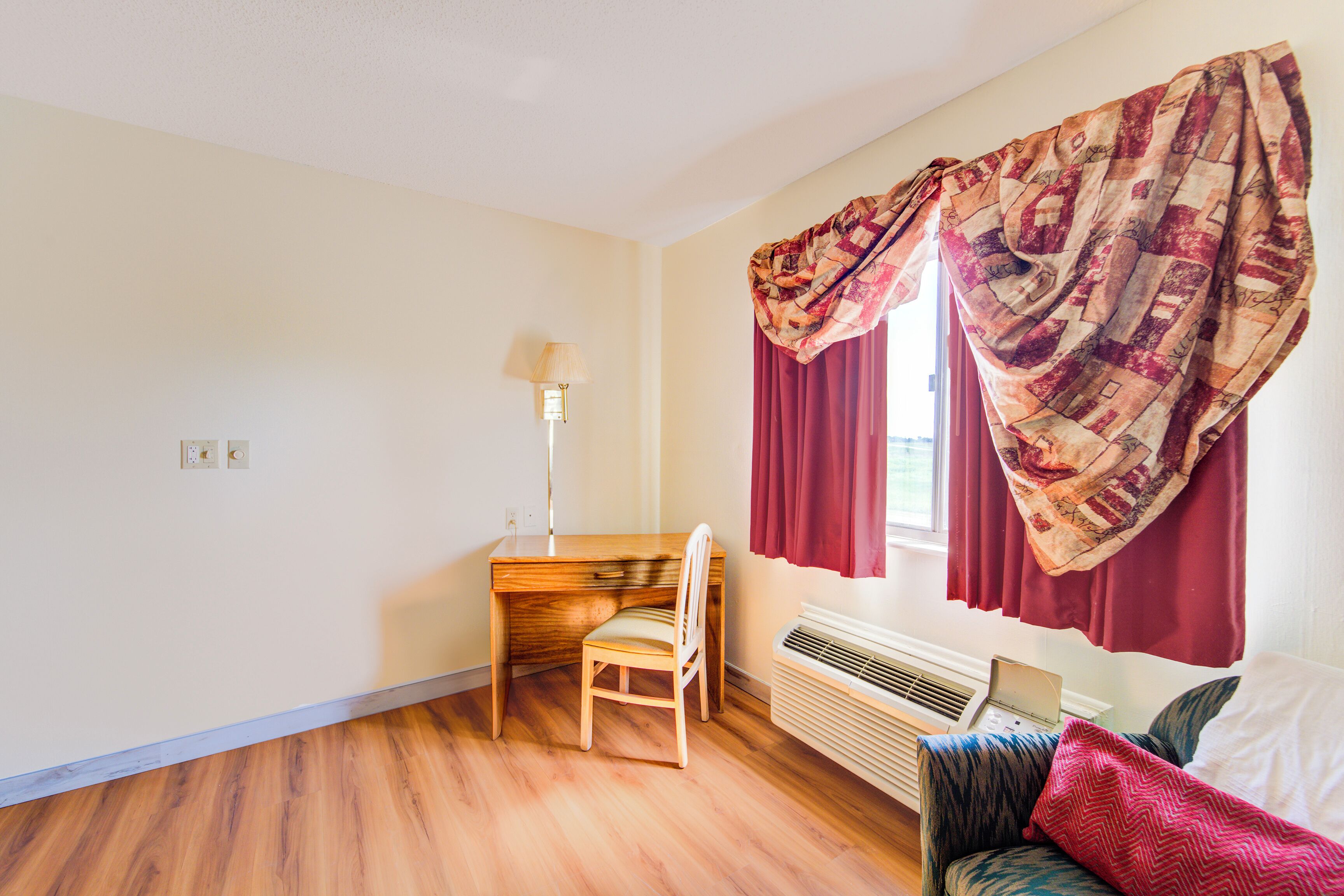 A room with a wooden desk, a chair, a lamp, a bed with patterned bedding, and a window with curtains.