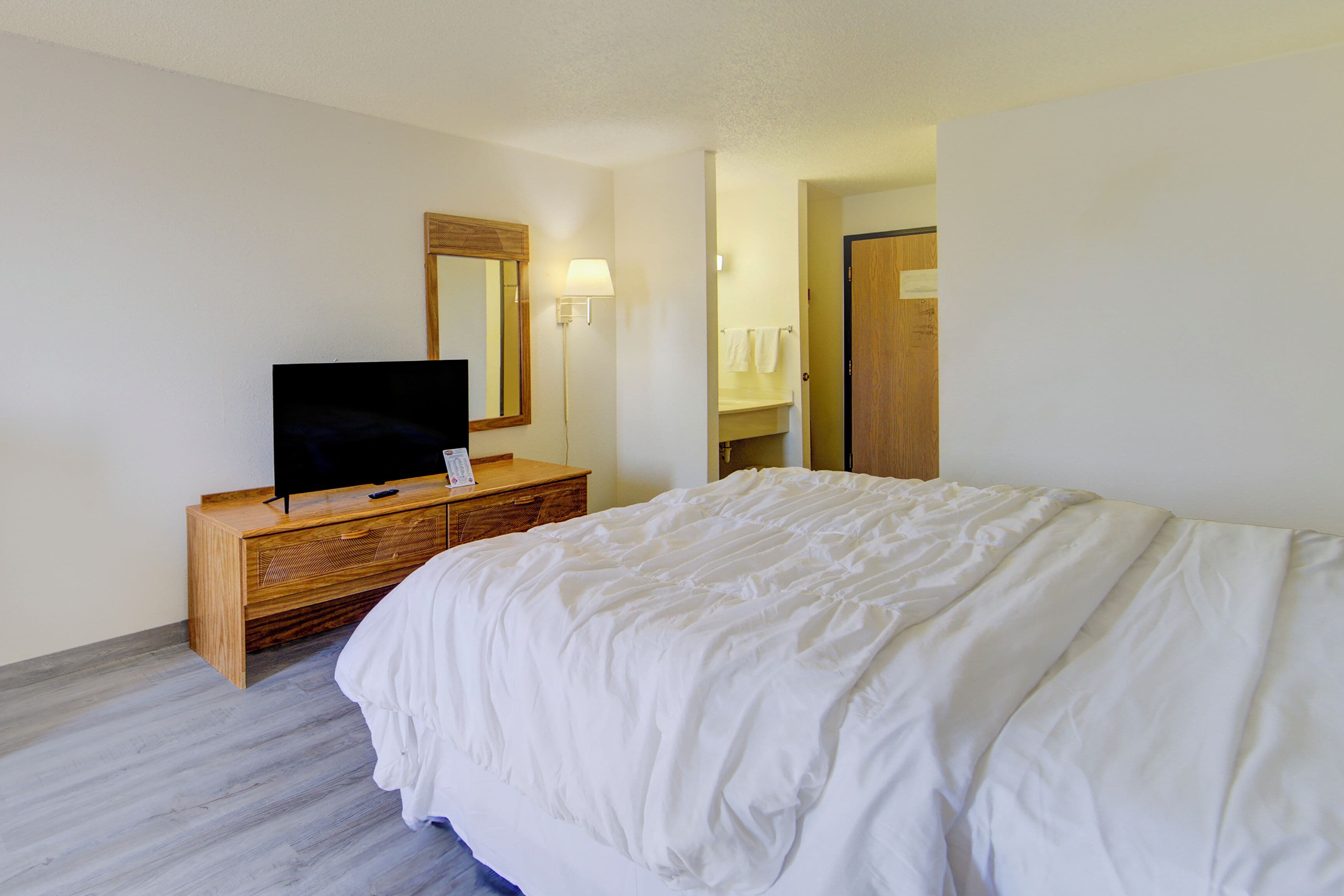 A hotel room with a bed, a TV on a wooden dresser, a mirror, and a bathroom visible through an open door.