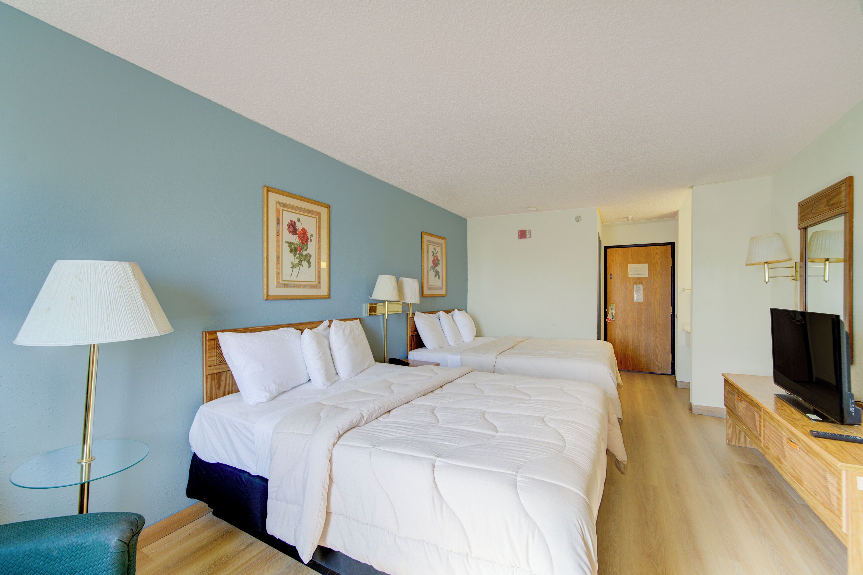 A hotel room with two beds, a television, a lamp, and framed pictures on the wall.