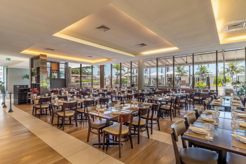 A spacious dining area with multiple tables and chairs, large windows offering views of palm trees, and a modern ceiling design.