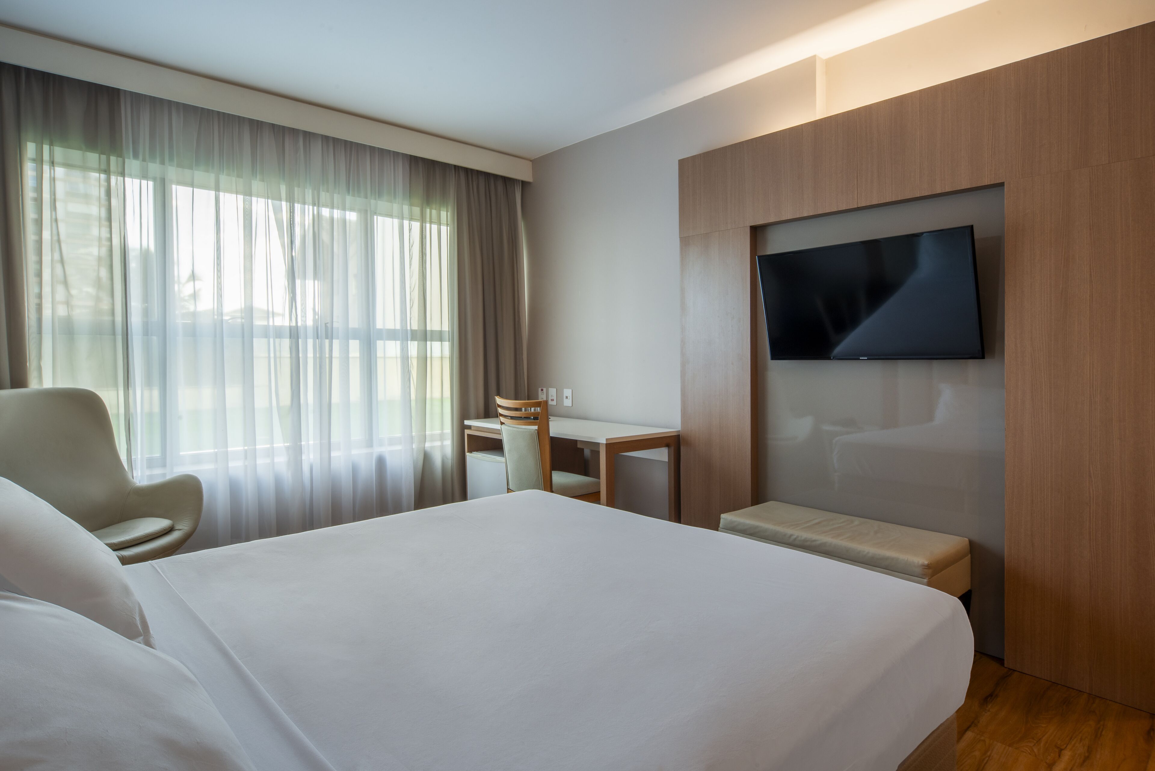 A hotel room with a large bed, a desk, a chair, a TV mounted on the wall, and a window with sheer curtains.