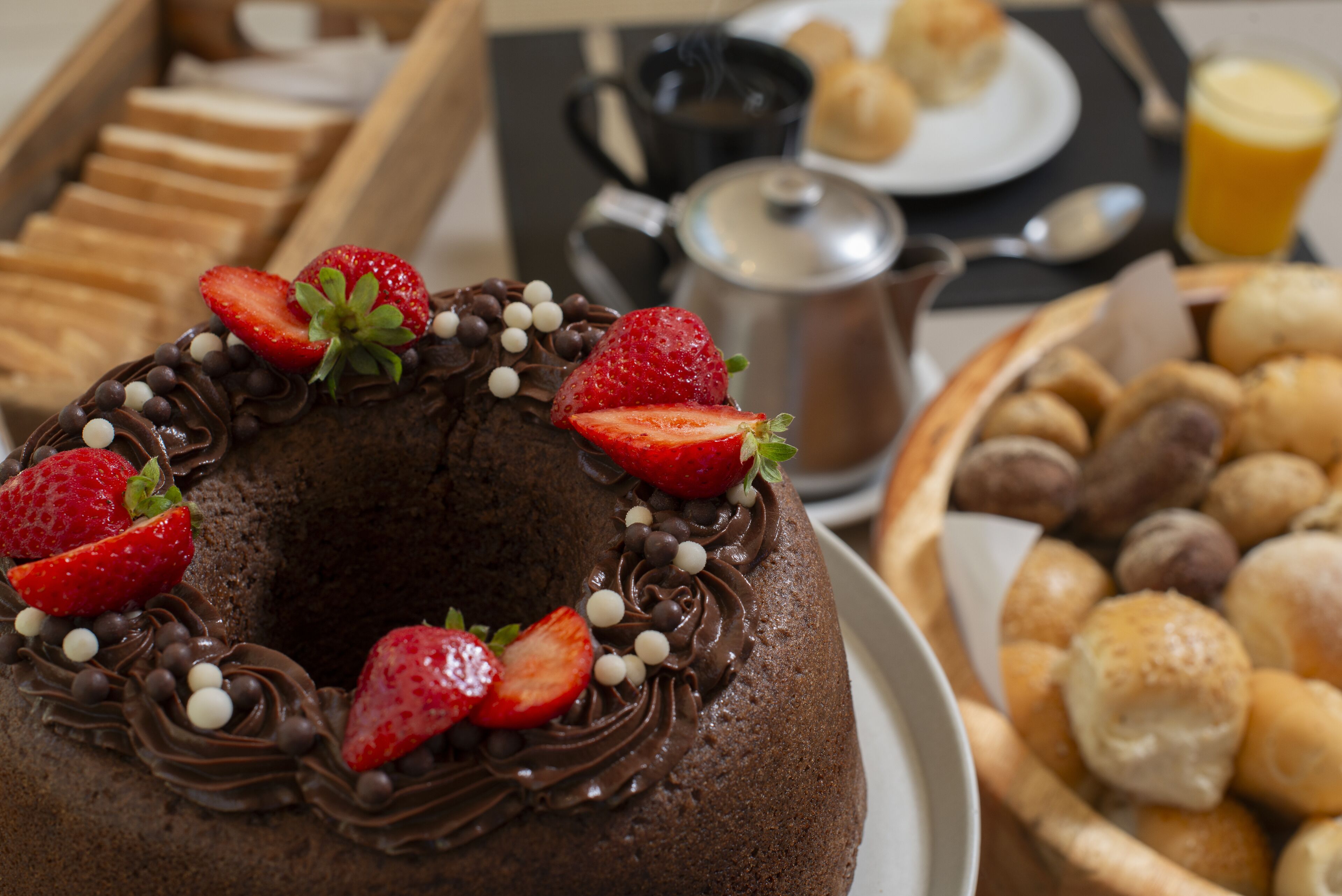 Un gâteau au chocolat garni de fraises et de pépites de chocolat, une cafetière et un panier de viennoiseries.