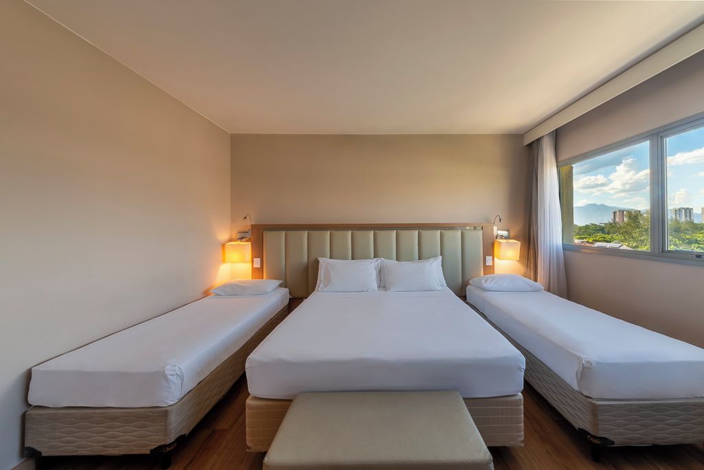 Une chambre d’hôtel avec trois lits, une tête de lit, des lampes de chevet et une fenêtre donnant sur un beau paysage.