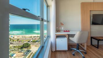 Habitación con un ventanal grande que da a una playa, un escritorio con silla, un refrigerador pequeño y un televisor montado.