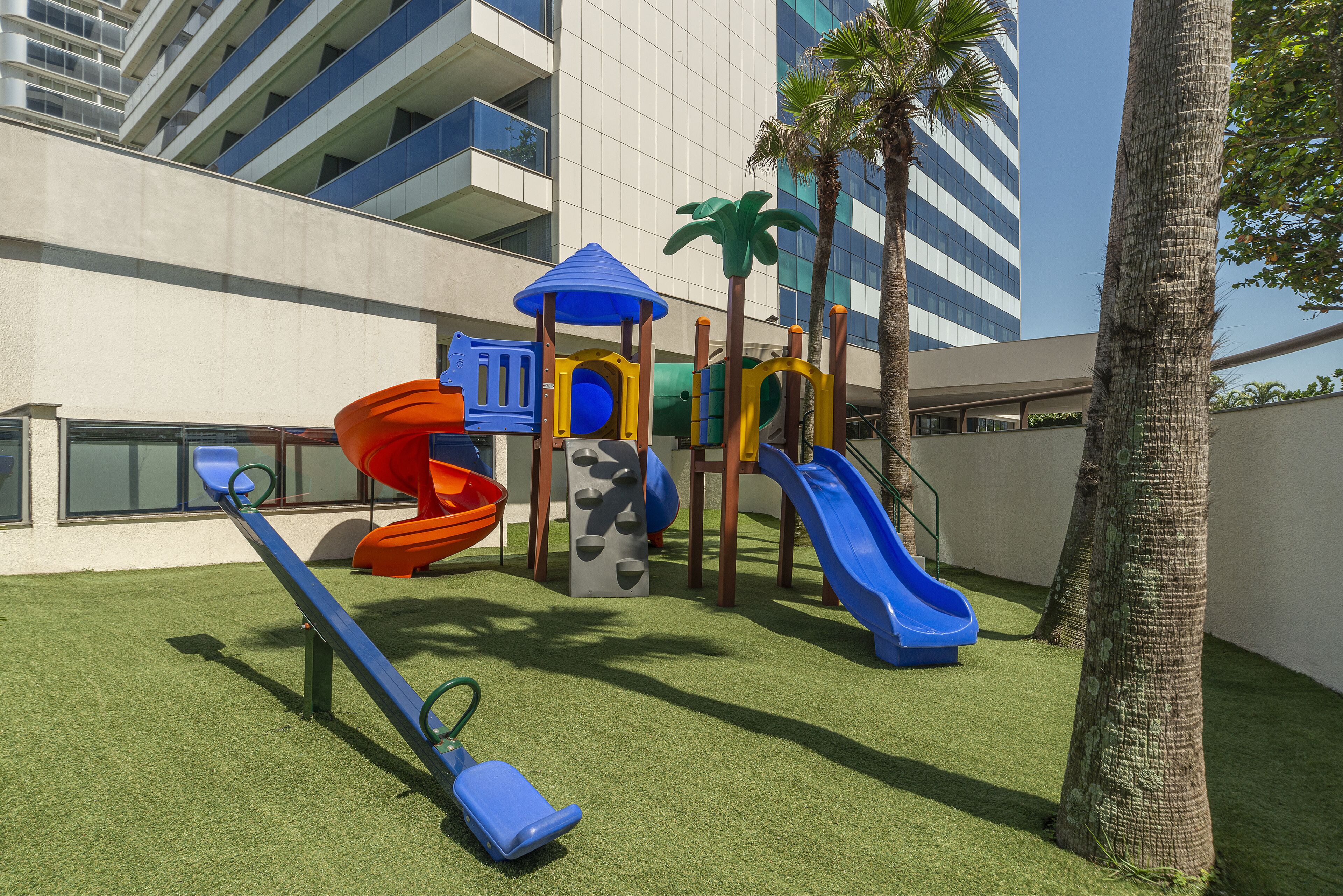 Un parque infantil con toboganes y estructuras para escalar, rodeado de palmeras y un edificio al fondo.