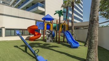 Un parque infantil con toboganes y estructuras para escalar, rodeado de palmeras y un edificio al fondo.