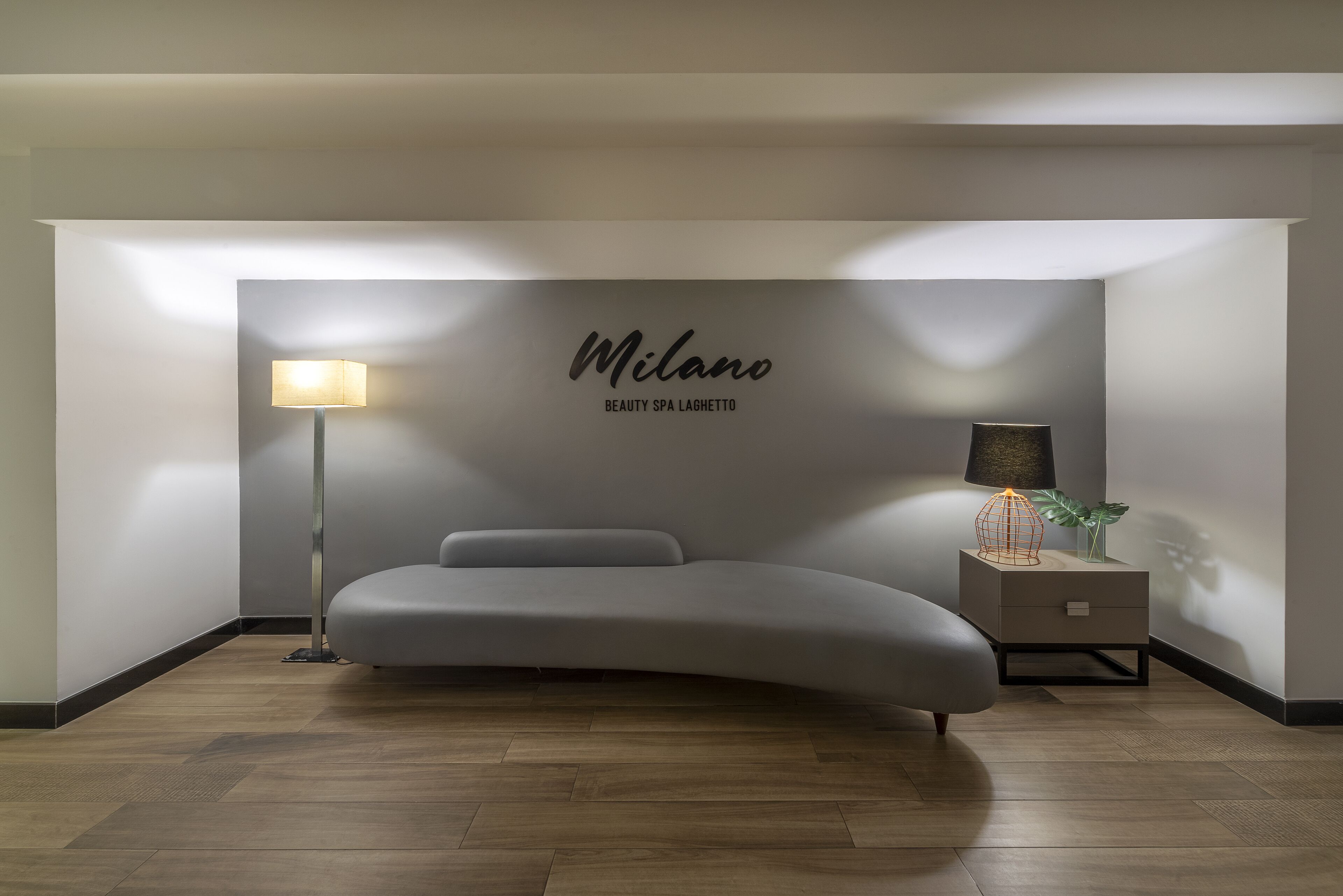 A modern bedroom with a curved bed, a bedside lamp, and a wall-mounted sign reading "Milano Beauty Spa Laghetto."