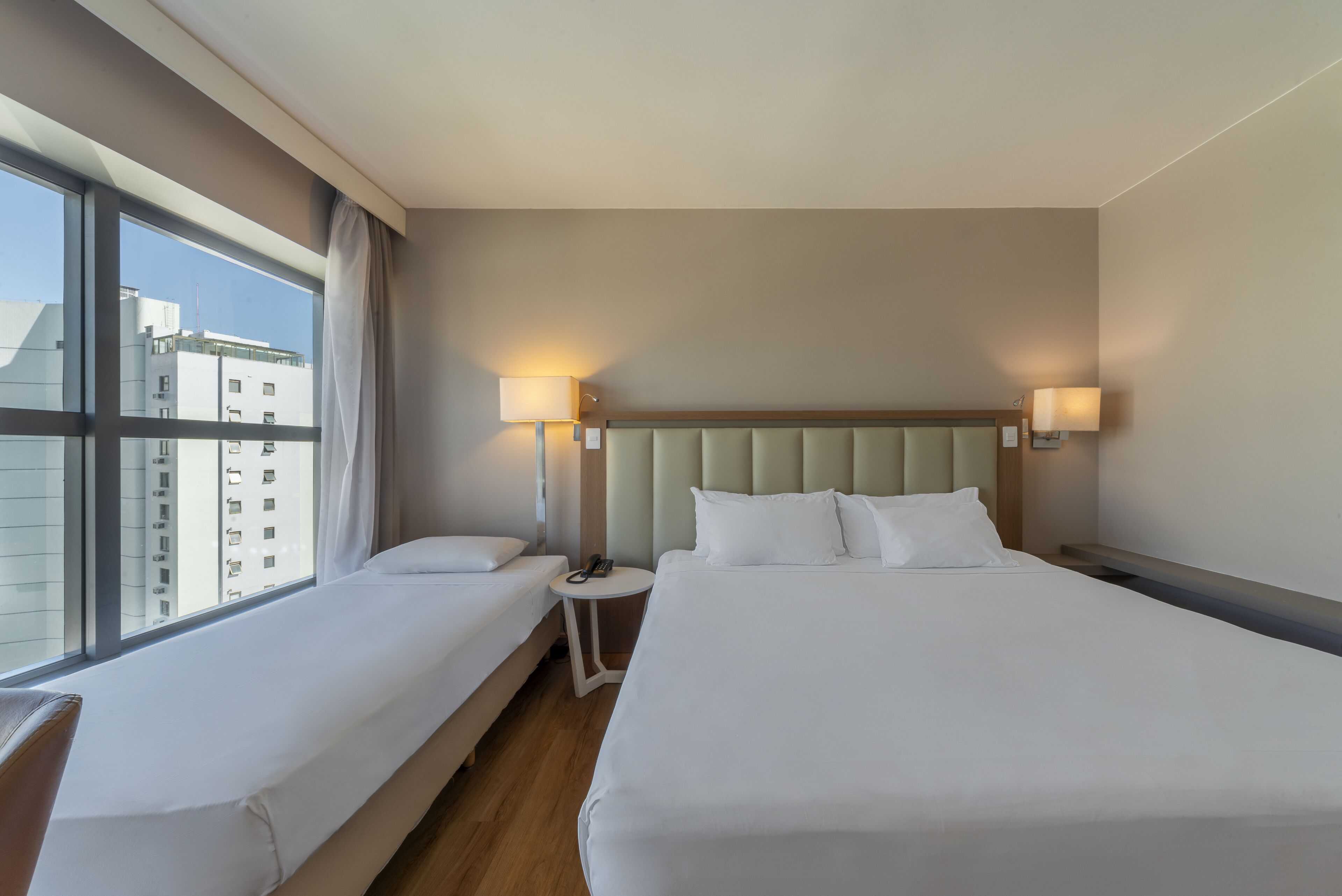 Habitación de hotel con dos camas, un ventanal grande y vistas a edificios.