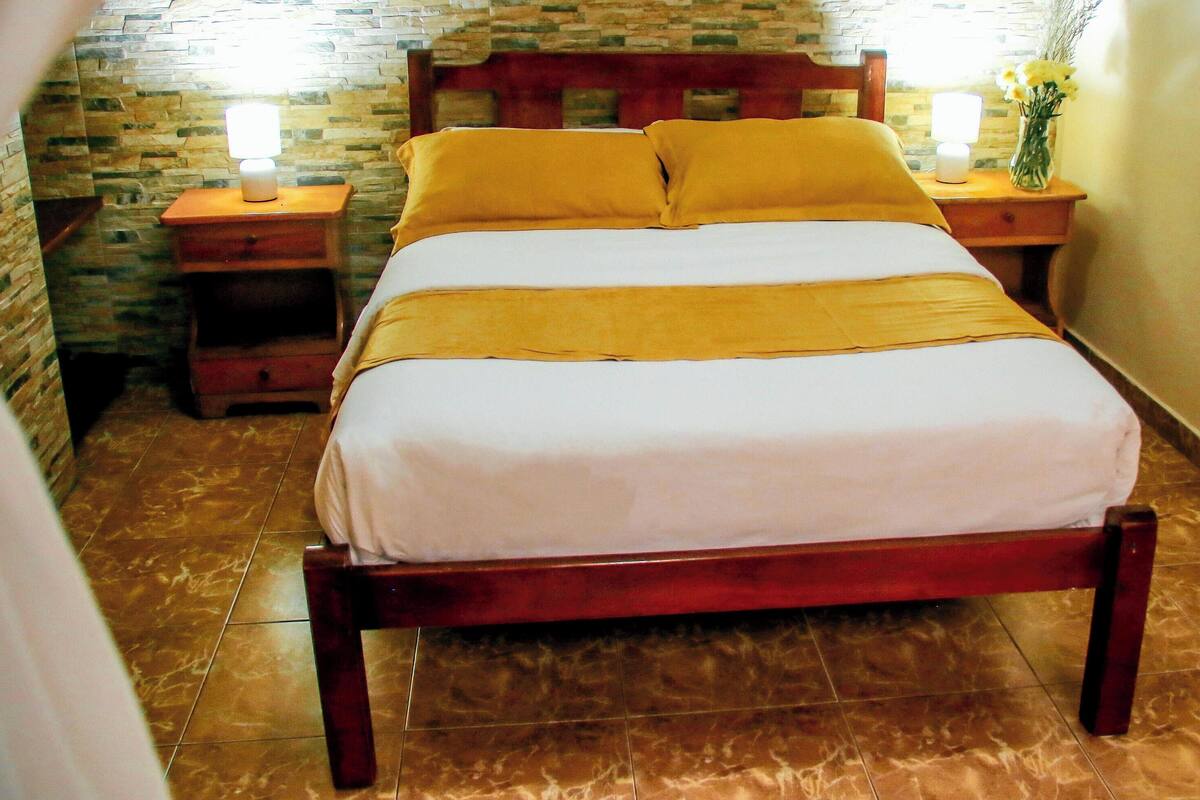 A bed with a wooden frame, a white mattress, and yellow bedding. There are two bedside tables with lamps. The room has a stone wall and a tiled floor.