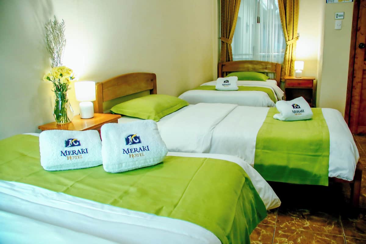 A hotel room with three beds, each with a green bedspread and a Meraki Hotel towel. There is a nightstand with a lamp and a vase of flowers.