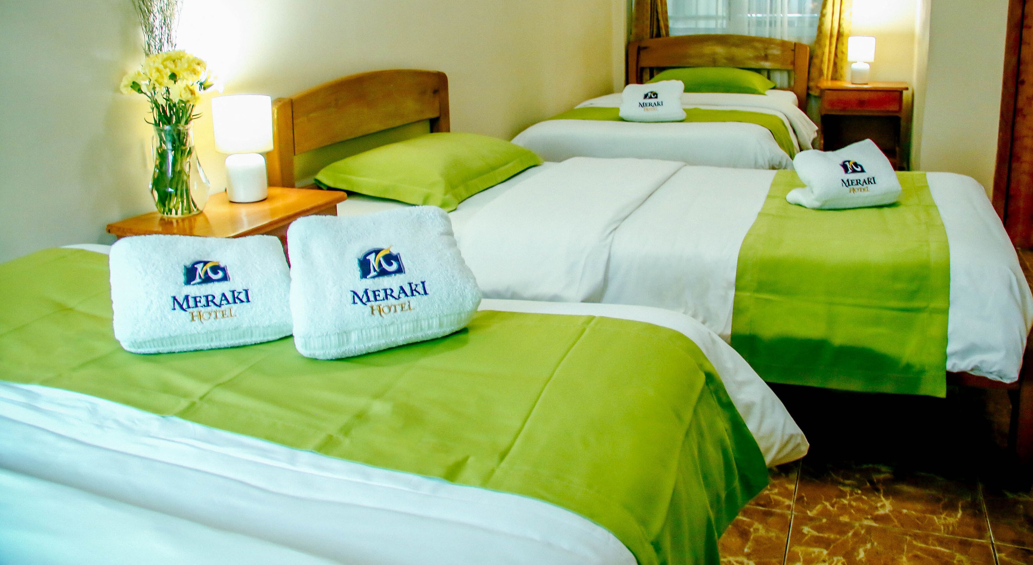 Habitación de hotel con tres camas, cada una con una colcha verde y un almohadón blanco con el logotipo del hotel. Hay una mesita de noche con una lámpara y un jarrón con flores.