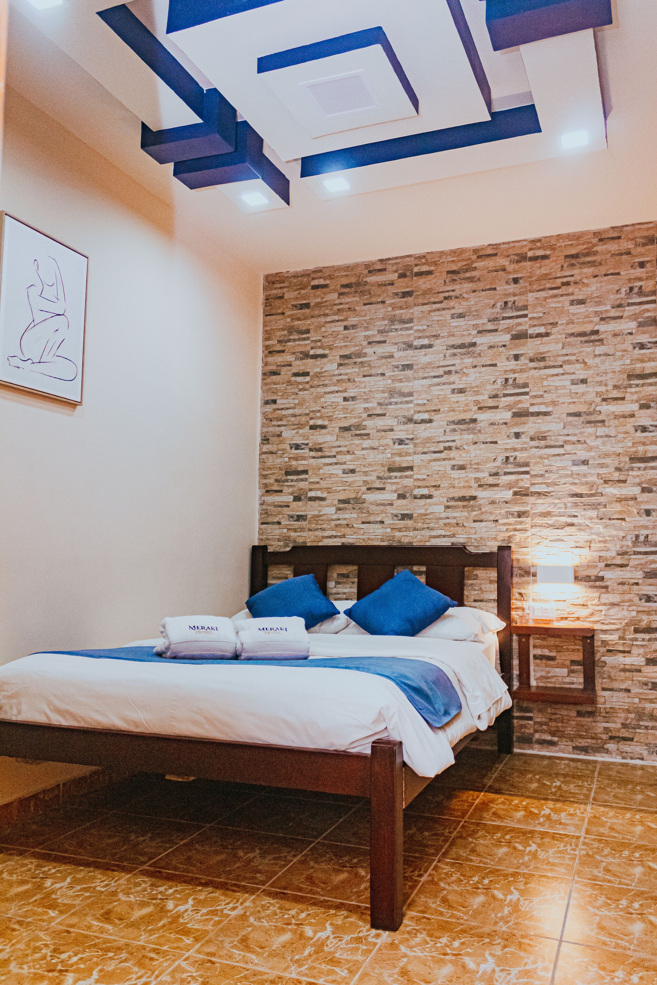 Een slaapkamer met een bed, een nachtkastje, een muur met een bakstenen patroon en een plafond met geometrische lampen.
